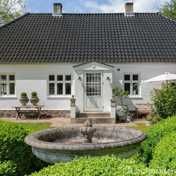 Et hvidt hus med sort tag står i en grøn have. Foran huset er der en rund stenfontæne omgivet af buske. En parasol er på terrassen ved husets højre side.