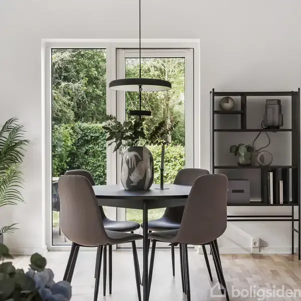 Et rundt spisebord med fire stole står i en moderne spisestue. Bordet har en vase med blomster. Bagved ses en reol ved siden af store vinduer med udsigt til en grøn have.