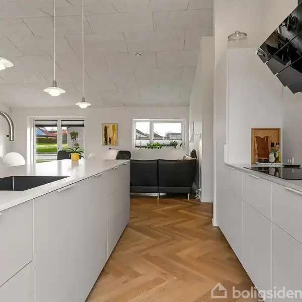 Køkken med hvide skabe og sort vask, imens pendellamper hænger over det. Til venstre er en sofagruppe ved vinduer, og trægulve dækker hele rummet.
