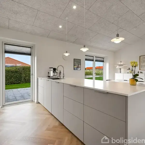 Et moderne køkken med en stor central ø, omgivet af lyse vægge og flere hængende lamper. Et spisebord med stole ses i baggrunden ved siden af store vinduer med udsigt til en have.
