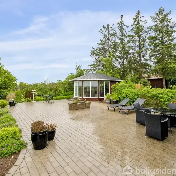 Havepavillon står på en stor terrasse omgivet af grønne planter og høje træer. Flere sorte havemøbler, inklusive stole og borde, er placeret strategisk omkring terrassen.