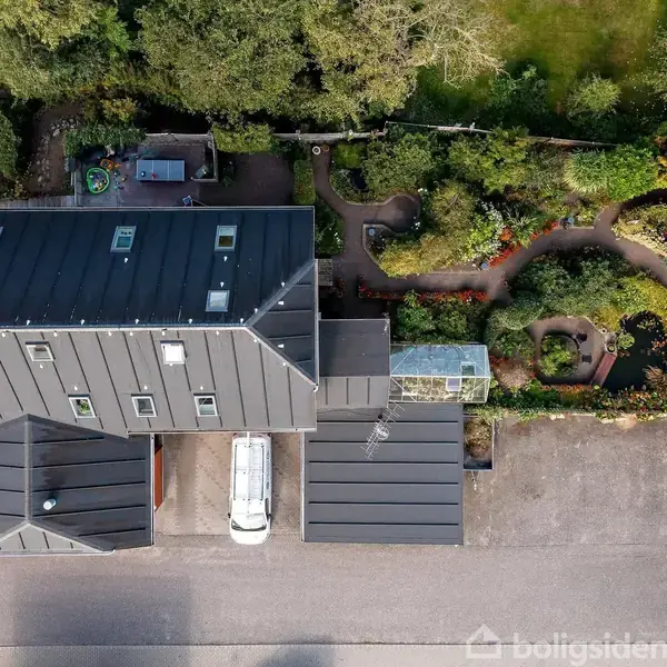 Hus set ovenfra, omgivet af grønne haver med stier og små planter. En glasstruktur er ved siden af huset. To biler er parkeret på grunden, en hvid og en rød.
