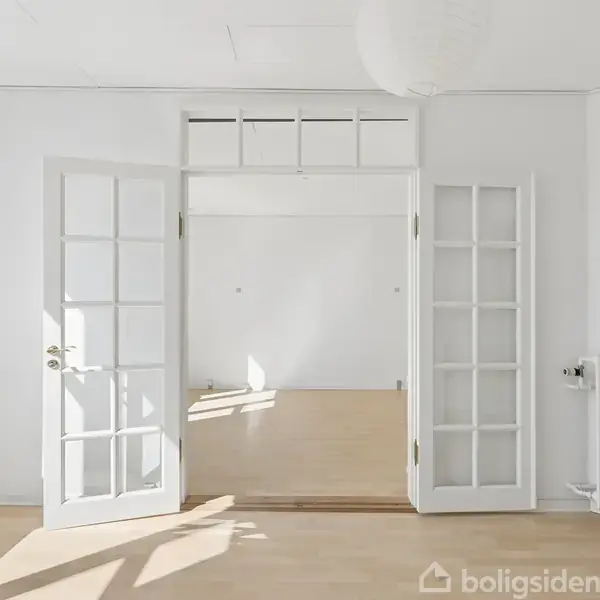 Dobbeltdør i glas står åben ind til et rummeligt, lyst rum med trægulv og hvidmalede vægge. Sollys strømmer ind gennem vinduerne og rammer gulvet, mens en radiator er synlig til højre.