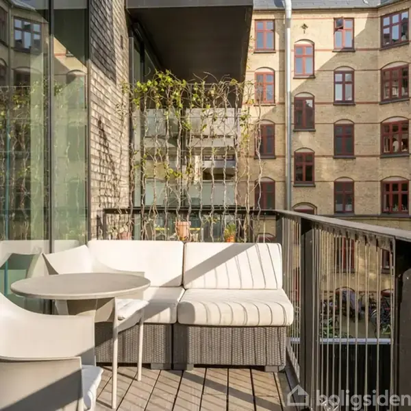 En balkon med en moderne sofa og et rundt bord med tilhørende stol. Balkonen vender mod en indre gård, omgivet af gamle murstensbygninger med røde vinduer.
