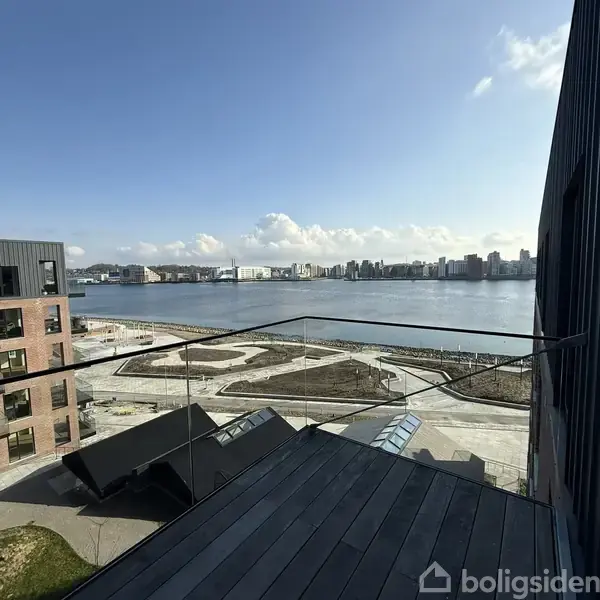 En altan med glasrækværk med udsigt over en bred flod. I baggrunden ses en by med moderne bygninger under en blå himmel med skyer.