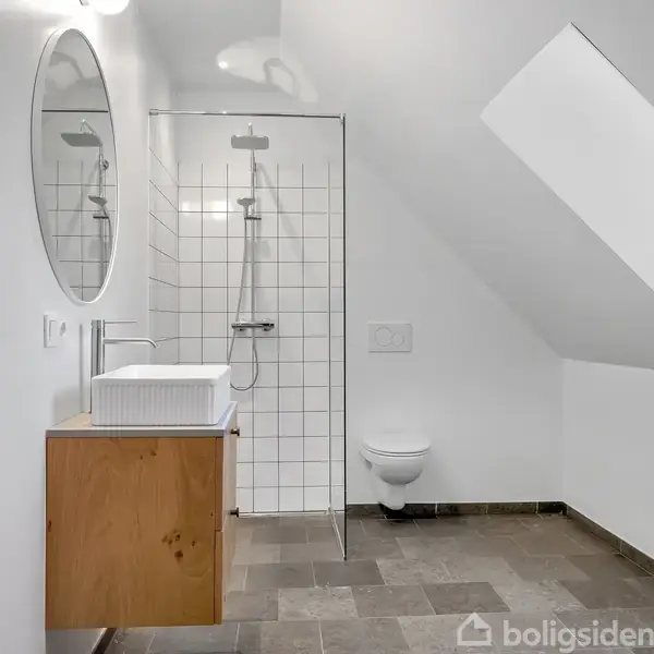 Brusekabine med glasvæg og hvidt flisebelagt baggrund. Til venstre er et træskab med vask og rund spejl. Værelset har et skråtag og et toilet ved siden af bruseren.