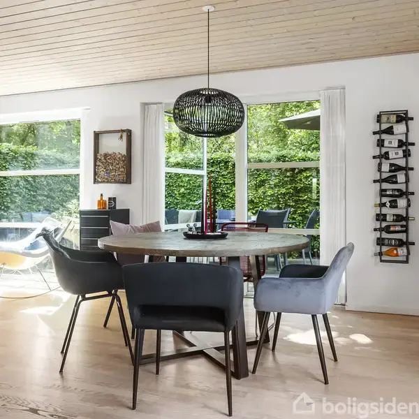 Rundt spisebord omgivet af moderne stole står i et lyst rum. Store vinduer giver udsigt til en frodig have, mens en vinreol og kunstværker dekorerer væggene.