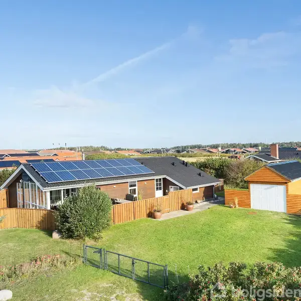 Et hus med solpaneler på taget står i en have med græsplæne, omgivet af hegn. En garage til højre. Omkringliggende nabolag med flere huse ses i baggrunden.