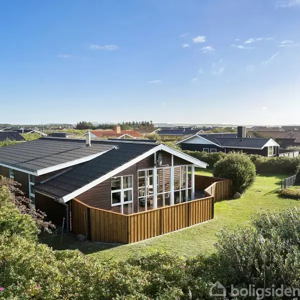 Et moderne sommerhus med store vinduer og skråt tag står i et grønt og frodigt landskab. Flere huse anes i baggrunden under en klar blå himmel.