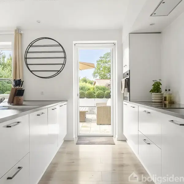 Et moderne køkken med hvide skabe og grå bordplader. Udendørs udsigt til terrasse gennem glasdør, omgivet af grønne planter. Rund sort væghylde og knivholder på bordet.