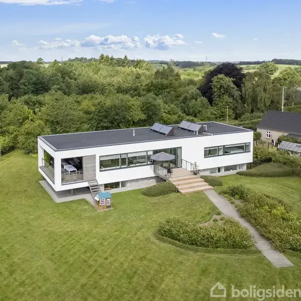 En moderne, hvid enfamiliesvilla ligger midt i en stor, velplejet græsplæne omgivet af skov og andre huse. Villaen har en flad tagkonstruktion og store vinduespartier.