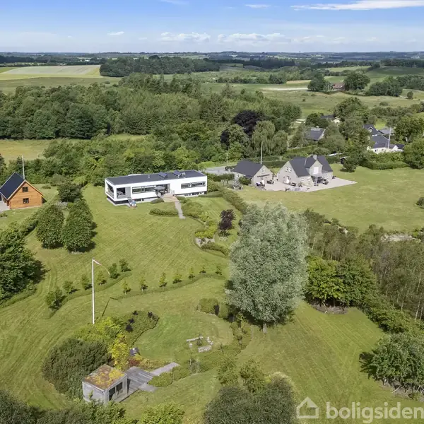 Et moderne, hvidt hus står på en stor grøn grund. Omgivet af træer og flere huse, med marker og skov i baggrunden under en blå himmel med få skyer.