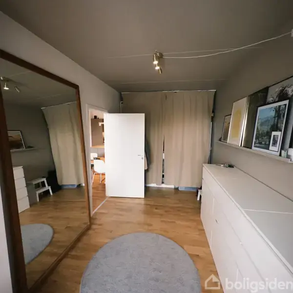 Et spejl står på et trægulv i et rum med en åben dør, som fører til et andet rum med en stol. Gardiner hænger foran et vindue, og der er skabe med billeder ovenpå.