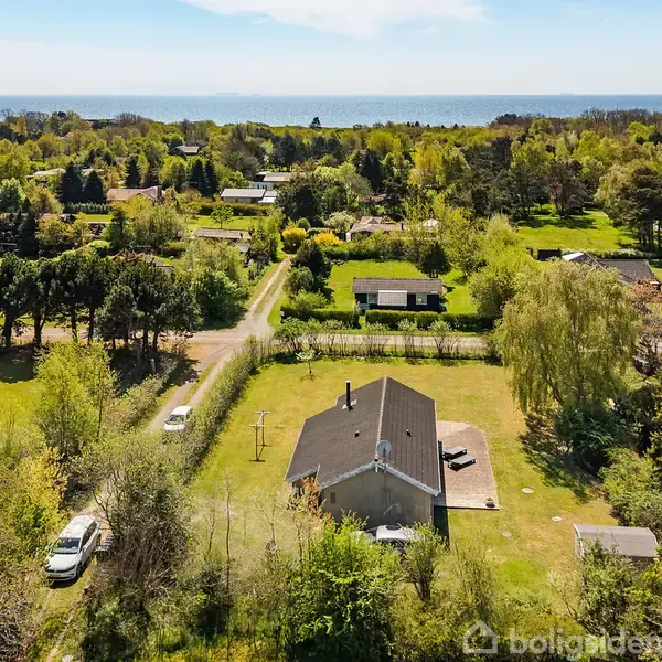 Hus set fra oven omgivet af grøn have og træer, med kystlinje i baggrunden. Flere huse og veje omkranser området, som er præget af frodig vegetation.