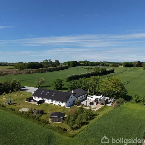 Hvid bygning med sort tag ligger i en landlig eng omgivet af grønne marker og træer under en blå himmel. Der er også en legeplads og en pavillon i nærheden.