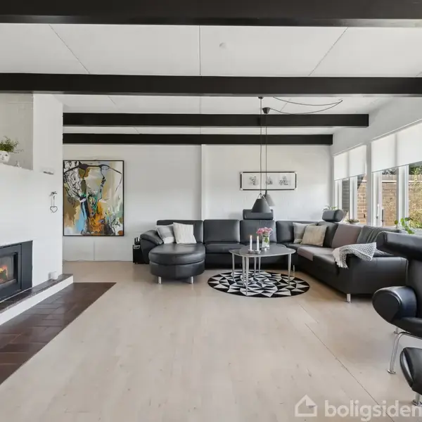 Stue med moderne, sorte lædersofaer og pejseindsats til venstre. Store vinduer lader dagslys ind, omkranset af hvide vægge og mørke loftsbjælker. Rundt sofabord med sort-hvidt tæppe.