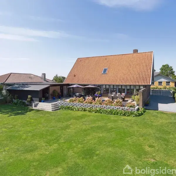 Et stort hus med rødt tegltag står omkranset af grønne plæner og hække. En terrasse med havemøbler og parasoller er foran huset. En blå himmel i baggrunden.