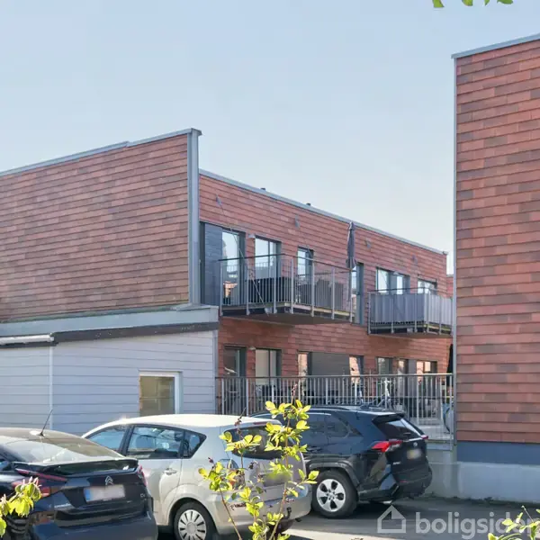 Moderne bygninger med røde facader står stille bagved parkerede biler. Sollys skinner på de store vinduer og altaner. Grønt løv rækker ind fra kanten af billedet i forgrunden.