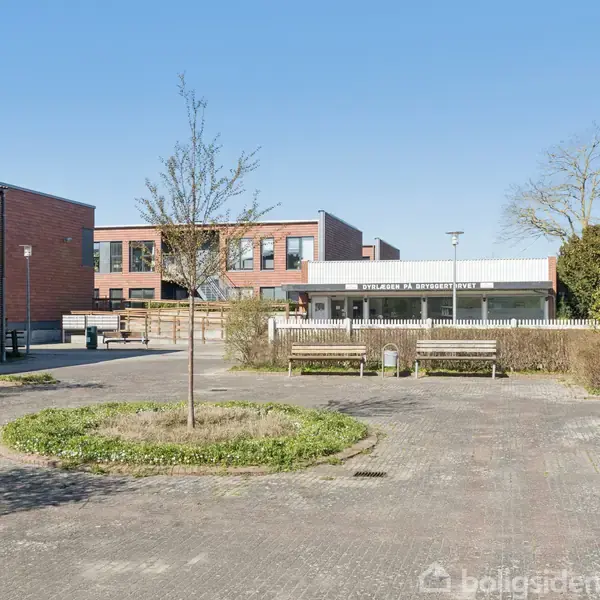 En åben gårdsplads med en bygning i baggrunden og omgivet af buske. Bænke står ved siden af græsarealer. En træ i midten af gårdspladsen.