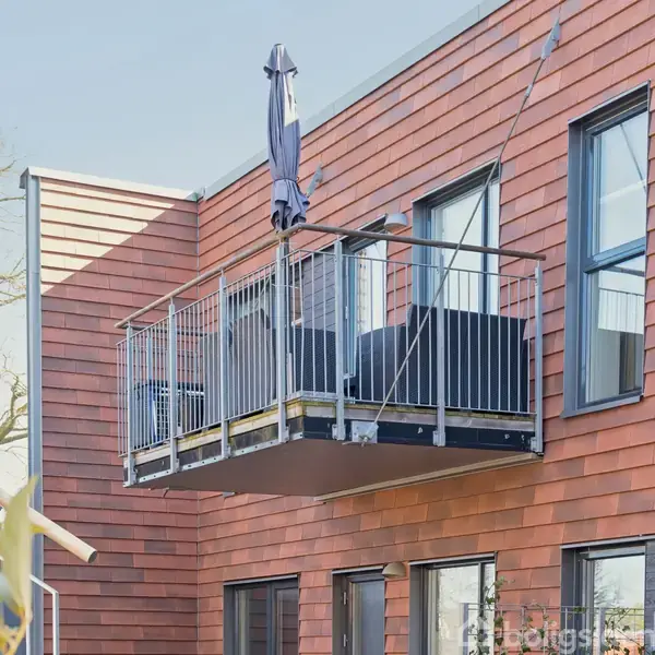 Balkon med møbler og parasol hænger ud fra en moderne rød murstensbygning. Ruderne reflekterer lyset, og der er træer i baggrunden.