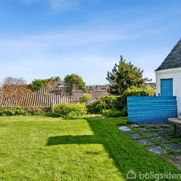Hus i solskin med hvid facade og blå vindue, står ved siden af grøn græsplæne med buske og træer i baggrunden omkranset af et hegn under en blå himmel.