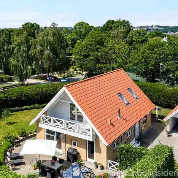 Et hus med rødt tag står omgivet af en frodig grøn have. Ved siden af huset er en carport. I baggrunden ses skovbevoksning og en sø i et landskab.