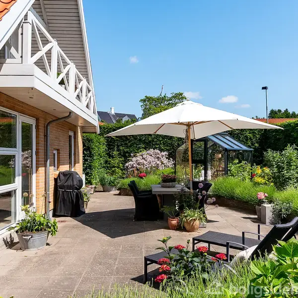 Terrasse med havemøbler og parasol foran et hus med rødt tag. Grill og blomsterkrukker står spredt på flisebelægningen. Hækkene omkranser området, og et væksthus ses i baggrunden.