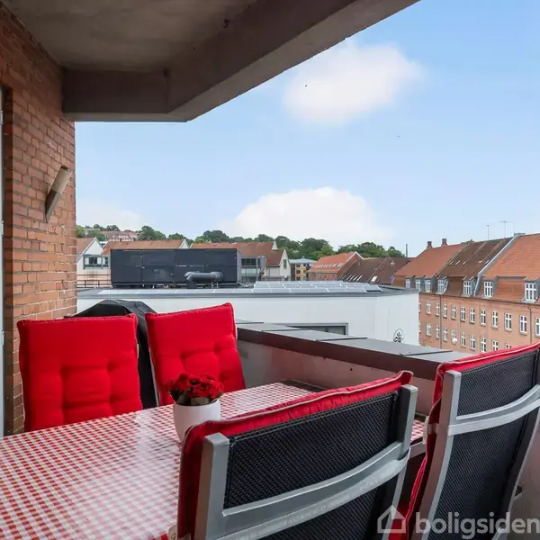 Balkon med røde polstrede stole omkring et bord med rød- og hvidternet dug, placeret ved murstensvæg. Udsigt over byens tage og en klar blå himmel.