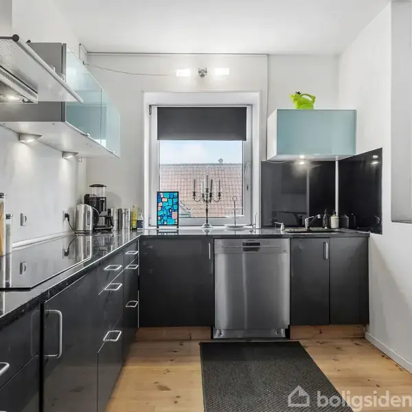 Et moderne køkken med sorte skabe og marmorbordplader. Køkkenet indeholder en opvaskemaskine, elkedel og andre køkkenredskaber. Et vindue over vasken giver udsigt til røde murstensbygninger.