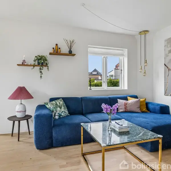 En blå sofa står i en stue ved et vindue, omgivet af planter og små hylder. En glasbord med blomster står foran sofaen. En lysestage hænger fra loftet.