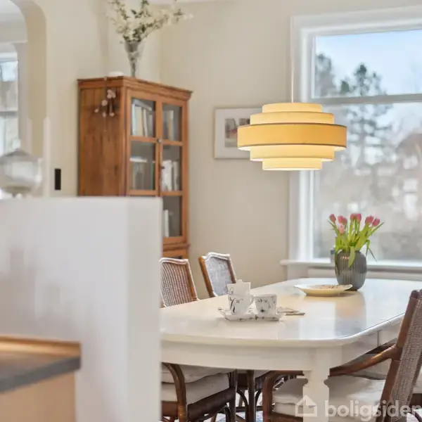 Et ovalt spisebord står dækket med kopper og tallerkener. Det er omgivet af stole og oplyst af en hængelampe. Et skab og vinduer med udsigt til træer udgør baggrunden.