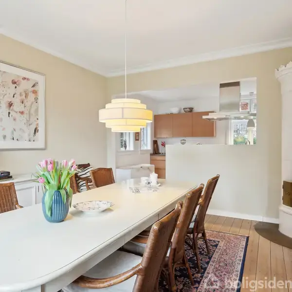 Spisebord med to vaser med blomster og fem stole står i et spisestueområde. En maleri hænger på væggen, og en gul lampe hænger over bordet. I baggrunden ses køkkenet.