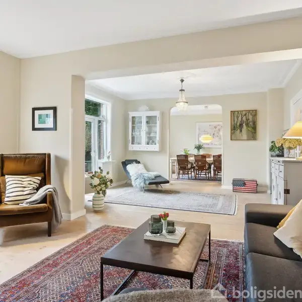 En stue med en grå sofa og lænestol, oplyst af lamper. I midten et sofabord. I baggrunden ses et spisebord i spisestuen. Væggene er cremefarvede.