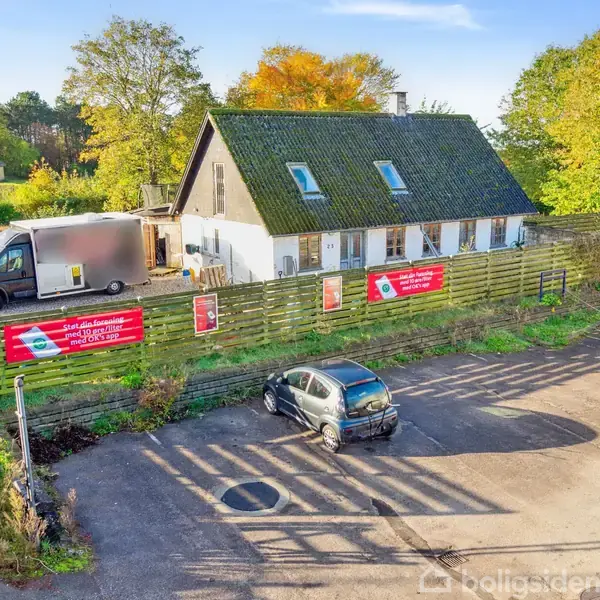 Hus med sort tag og grøn træhegn omkranset af to biler, en varevogn og en personbil, parkeret på asfaltplads. Skilte på hegnet: "Mexicansk mad i særklasse" med menuoversigt.