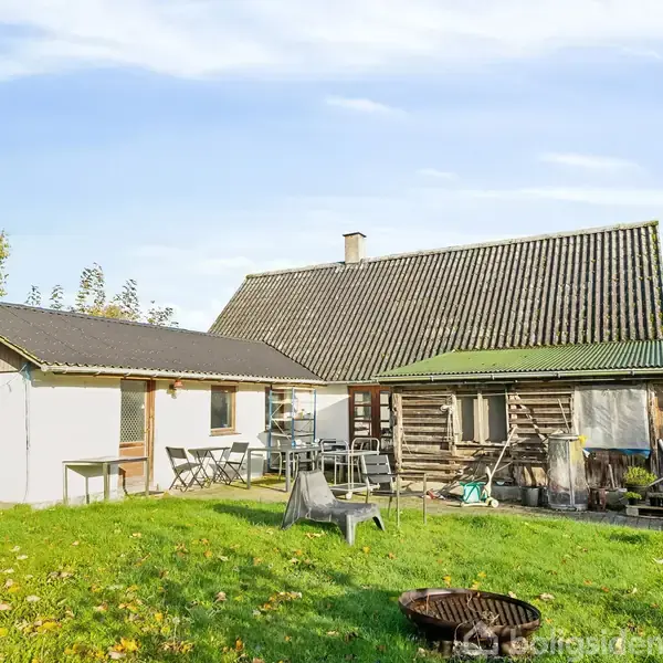 Et hus med to forskellige tagkonstruktioner, hvor et siddeområde med stole og borde er placeret i haven. Omkring ligger grøn græsplæne og træer under en blå himmel.