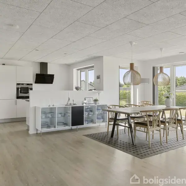 Moderne køkken-alrum med spisebord og seks stole. Store vinduer giver udsigt til have. Køkkenarea inkluderer ovn og emhætte. Lyse trægulve og loftslamper fuldender det minimalistiske look.