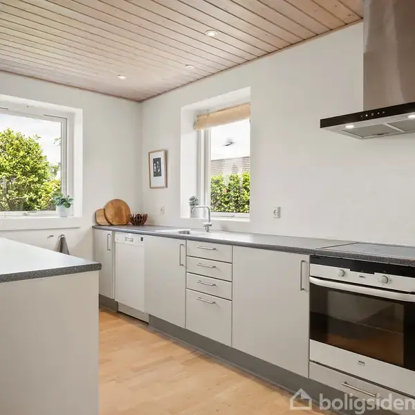 Et lyst, moderne køkken med hvide skabe og sort bordplade, hvor komfuret og emhætten er synlige. To vinduer giver udsigt til grønne buske udenfor.