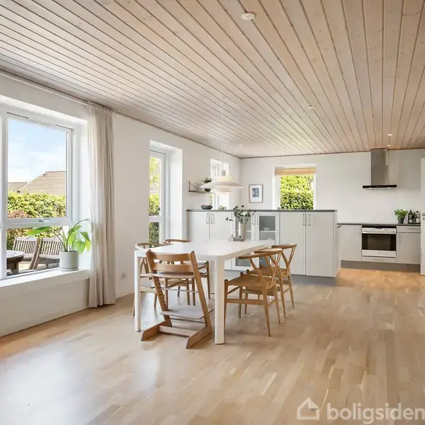 Et lyst køkken-alrum med spisebord og fire træstole. Rummet har lyse trægulve, hvidt køkken med moderne apparater. Store vinduer giver udsigt til en terrasse og grønne omgivelser.