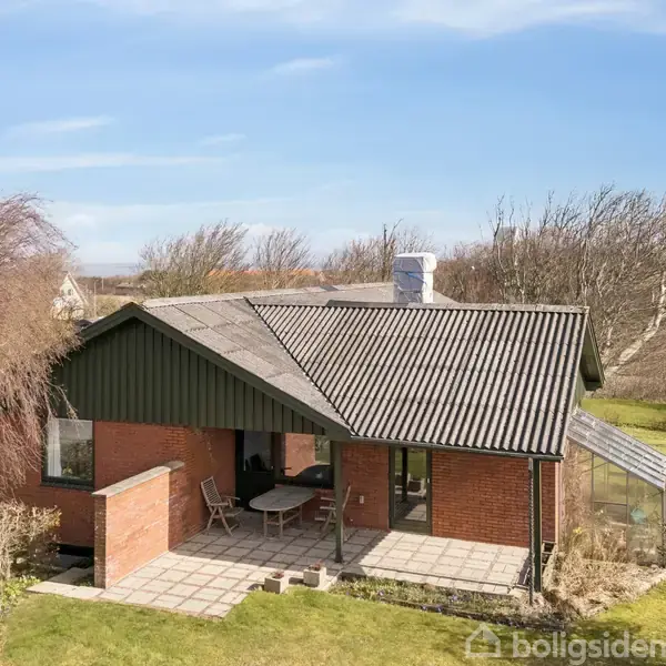 Et rødt murstenshus med gråt tag, stående i en have. Huset har en flisebelagt terrasse med stole. Træer omgiver bygningen, og himlen er blå med enkelte skyer.