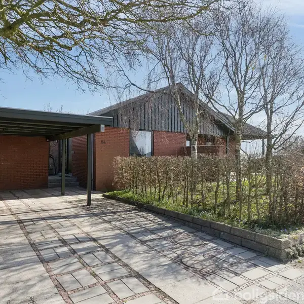 Hus står stille med røde mursten og fladt tag, omkranset af hække og træer. Til venstre ses en carport med flisebelagt indkørsel.