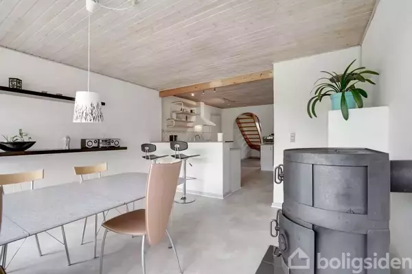 En moderne spisestue med et rundt bord og stole, en ovn i forgrunden. Køkkenbaren med to barstole er synlig i baggrunden. Væggene har hylder og en hængende lampe.