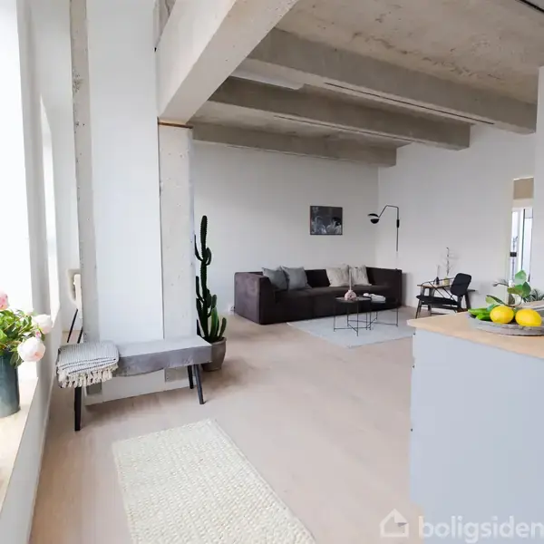 Stue med sofa og bord, omkranset af lyse vægge og store vinduer. Blomster og frugter pynter rummet, der har en minimalistisk indretning med betonloft.