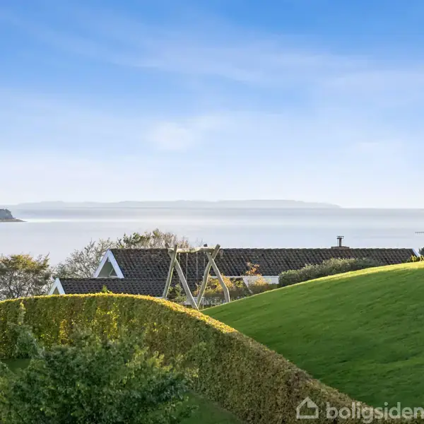 Et grønt, skrånende landskab med hække omkredser en bolig beliggende nær kysten. I baggrunden ses havet og en ø ved horisonten under en blå himmel.