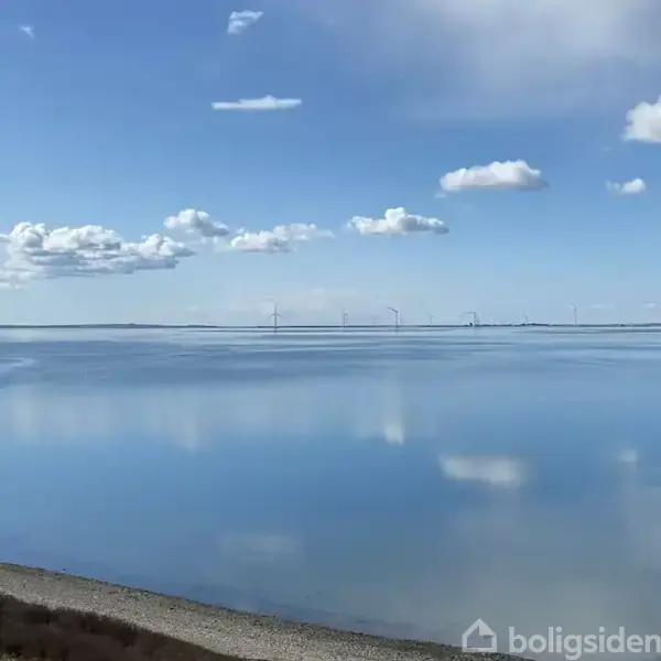 Vindmøller står stille på en rolig havoverflade under en næsten skyfri himmel med få hvide skyer, der spejler sig i vandet.