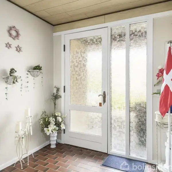 Dør entré. Hvid dør med glasruder, planter og blomsterdekorationer. Dansk flag står i hjørnet ved siden af et opbevaringsmøbel. Murstensgulv og lys væg med hængte dekorationer.