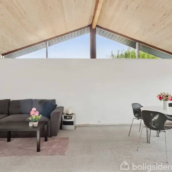 Sofa står i stuehjørnet med et kaffebord foran, mens et spisebord med fire stole er til højre. Store vinduer øverst giver udsigt til en blå himmel og grønt.