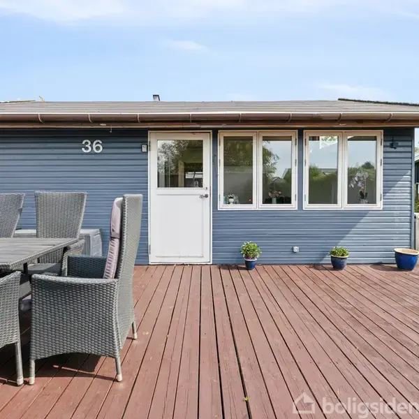Et sommerhus med nummer 36 vises med en stor træterrasse foran. På terrassen står et havebord med kurvestole. Huset er omgivet af en græsplæne og buske.