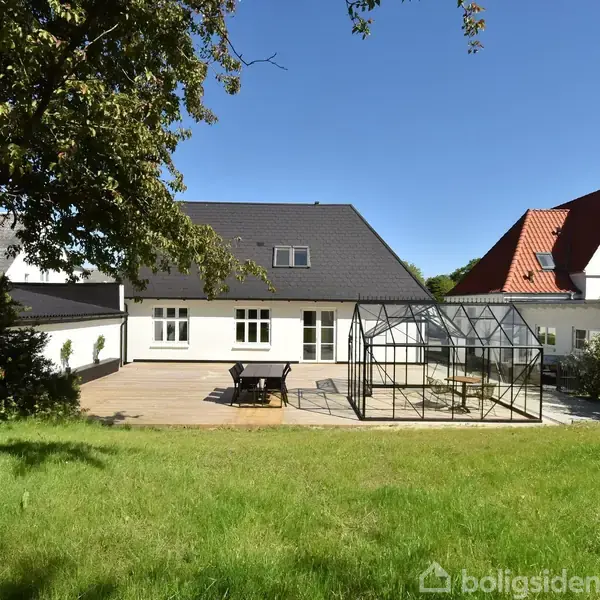 Hus med rødt tag og tilstødende drivhus på en stor terrasse. En træbord-bænk sæt står ved siden af drivhuset. Haven er grøn med græs og et stort træ til venstre.