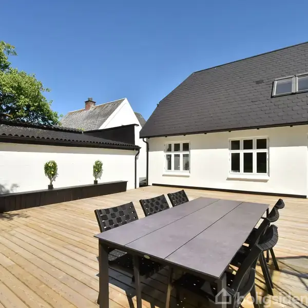 Et bord med stole er placeret på en stor træterrasse foran et hvidt hus med sorte tagsten. Der er to vinduer og en dør, omkranset af en have.