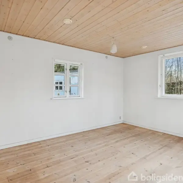 Køkken med hvide skabe og brunlige håndtag. En ovn står ved siden af en opvaskemaskine. Et vindue og en dør leder til en udendørs scene med træer.